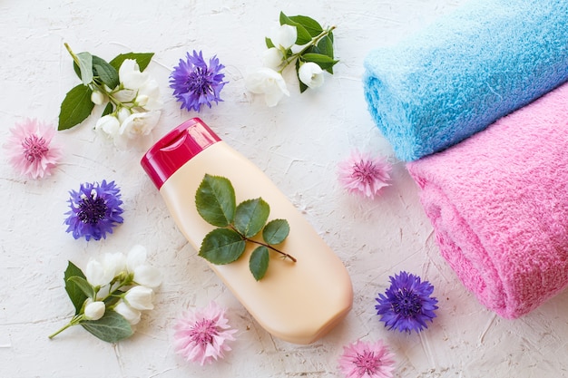 Flasche Shampoo, Handtücher und Blumen von Jasmin und Kornblume auf einer weißen Oberfläche