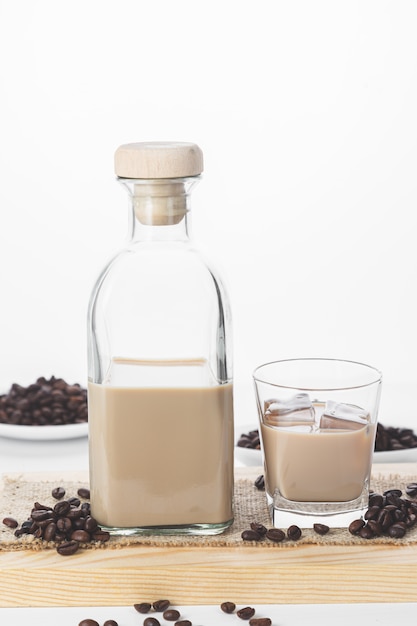 Flasche und Glas Sahne-Kaffeelikör mit Eis und Kaffeebohnen auf weißer rustikaler Oberfläche.