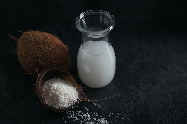 Flasche vegane Kokosmilch mit Streuseln, ganzer Kokosnuss und Flocken auf einem schwarzen Tisch. Gesundes Lebensstilkonzept.