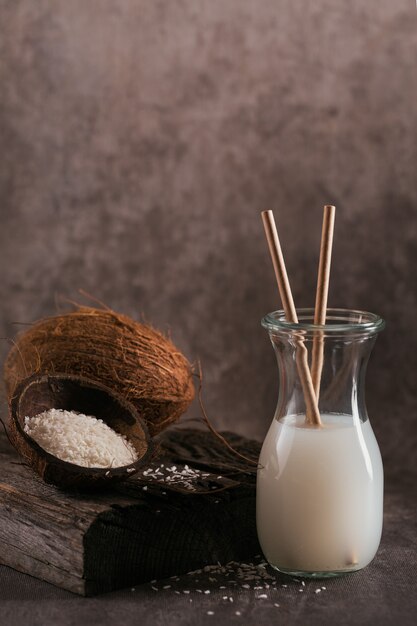 Flasche vegane Kokosmilch mit Strohhalmen, ganzer Kokosnuss und Flocken auf einem dunklen Tisch. Gesundes Lebensstilkonzept.