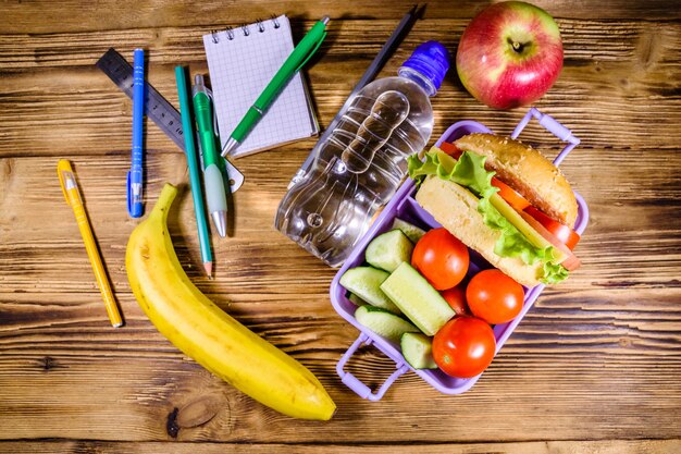 Flasche Wasser Banane reifer Apfel verschiedene Schreibwaren und Brotdose mit Hamburger Gurken und Tomaten auf rustikalem Holztisch Draufsicht