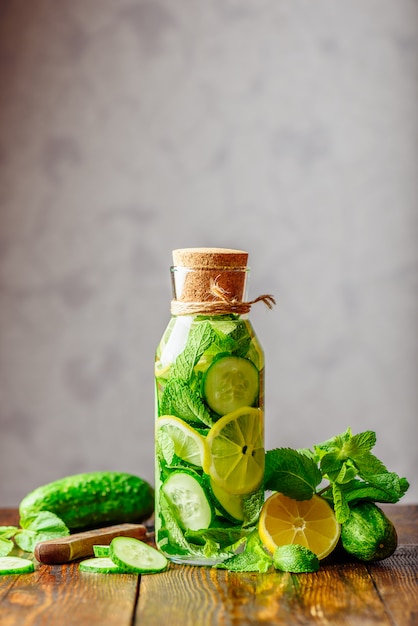 Flasche Wasser mit geschnittenen Zitronen-, Gurken- und Minzblättern. Zutaten und Messer auf dem Tisch.