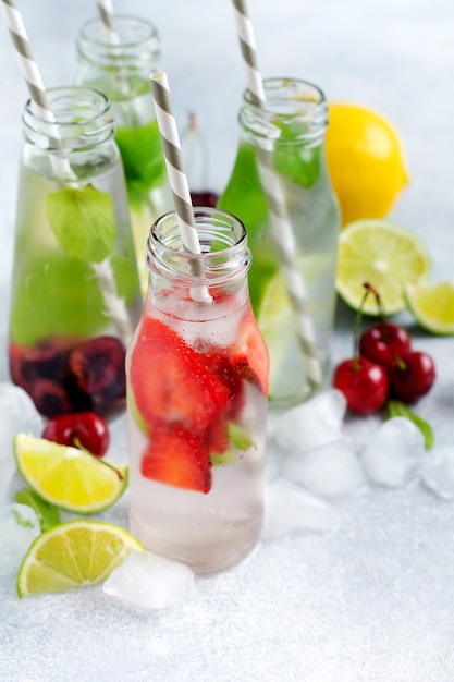 Flaschen Limonade erfrischende Sommerlimonade mit Limette, Erdbeere, Kirsche, Gurke und Eis auf einer grauen Betonoberfläche.