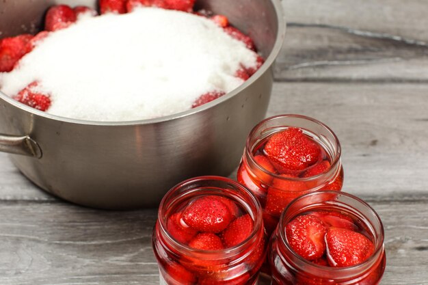 Flaschen mit hausgemachten eingelegten Erdbeeren, Stahltopf mit mehr mit Zucker bedeckten Beeren im Hintergrund. Hausgemachte eingelegte Fruchtzubereitung.
