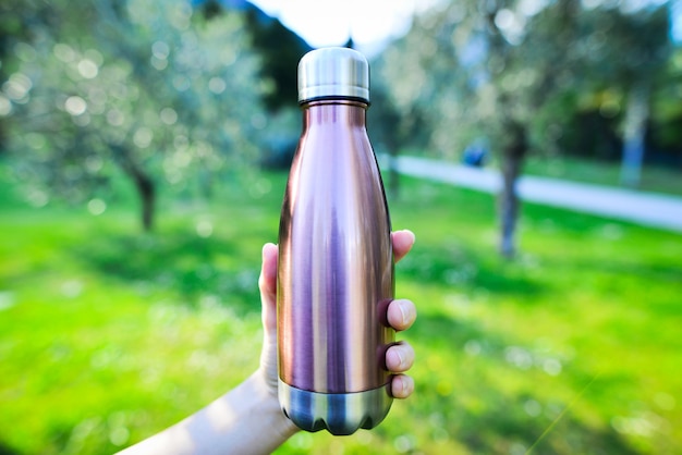 Flaschenwasser wiederverwendbar Nahaufnahme von Stahl-Eco-Thermowasserflasche in weiblicher Hand Aluminiumflaschenwasser wiederverwendbar auf dem Hintergrund Olivenbaumzweig mit unscharfer verschwommener umweltfreundlicher Wasserflasche