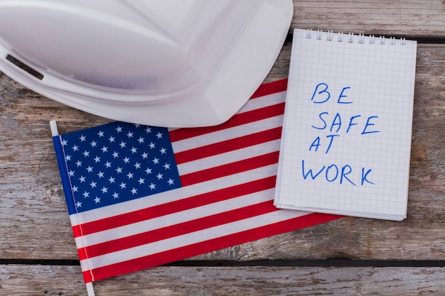 Foto flat lay warnt amerikanische bauarbeiter, um bei der arbeit sicher zu sein. weißer helm mit flagge der usa und notizblock auf gealtertem holztisch.