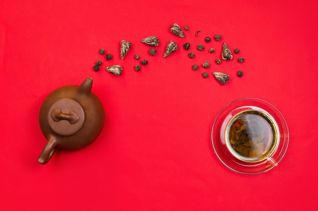 Flatlay-Anordnung mit chinesischer Ton-Teekanne und grünem Tee, die in eine Glasschale fallen. Roter Hintergrund.