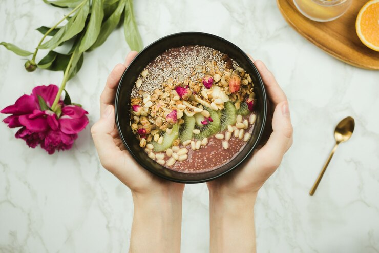 Foto flatlay das mãos da mulher segurando a tigela de batido vegan com pudim de chia coberto com granola, kiwi, pinhões e botões de rosa com flor de peônia e colher na mesa de mármore