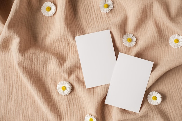 Flatlay de modelo de convite com estilo parisiense estético cartão de folha de papel em branco com espaço de cópia de maquete botões de flores de camomila em pano de musselina bege neutro vista superior plana