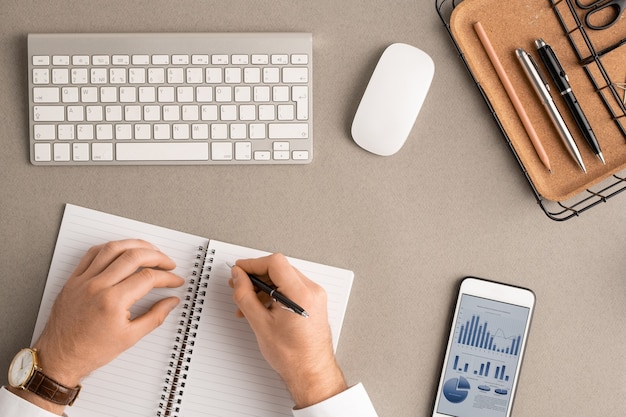 Flatlay der Hände eines jungen männlichen Brokers mit Stift über eine leere Seite eines offenen Notizbuchs, das Notizen zwischen Büromaterial, Maus, Gadget und Tastatur macht