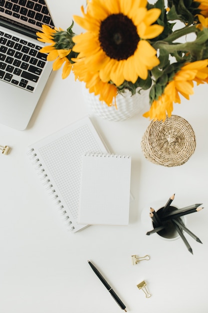 Flatlay del espacio de trabajo del escritorio de la oficina en casa con laptop, notebook, ramo de girasoles amarillos sobre blanco