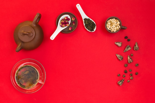 Flatlay-Rahmenanordnung mit chinesischen grünen Teeblättern, Rosenknospen, Jasminblüten und einer Teekanne aus Ton. Roter Hintergrund