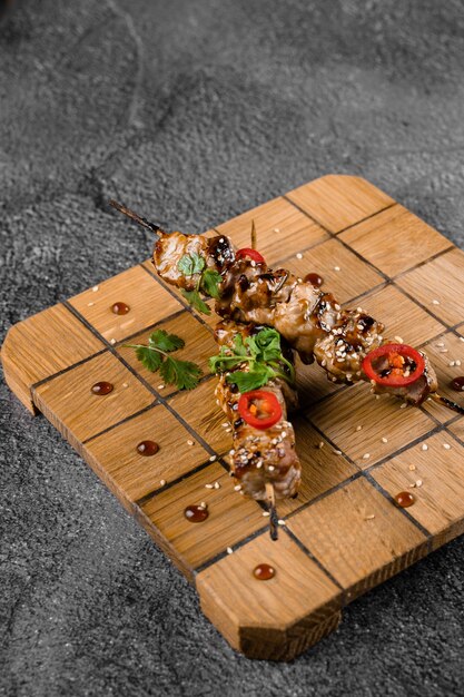 Fleisch auf Holzspießen, garniert mit Koriander, Sesam, Chili auf quadratischem Brett auf grauer Oberfläche.