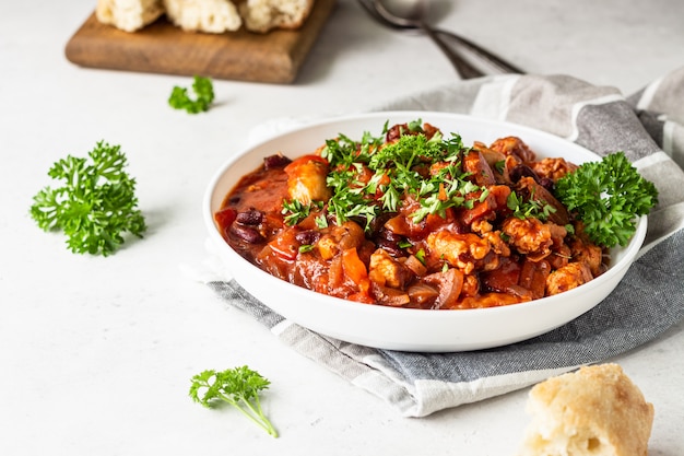 Fleischeintopf mit roten Bohnen, Paprika und Zwiebeln in Tomatensauce in einer weißen Platte über hellgrauem Schiefer