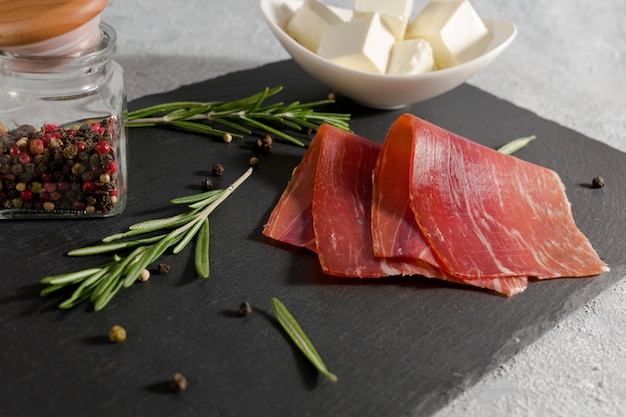 Fleischplatte, dünne Scheiben Schinken oder spanischer Jamon mit Rosmarinzweigen, Pfefferkörnern und eine Platte mit Weichkäse auf Steinschneidebrett