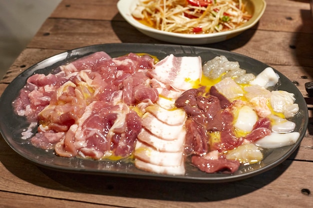 Fleischset für Sukiyakishabuyakiniku-Grill in einer schwarzen Platte auf einem Holztisch Das rohe Fleischset umfasst Rindfleisch, Schweinefleisch, Speck, Leberfisch, Tintenfisch und Garnelen