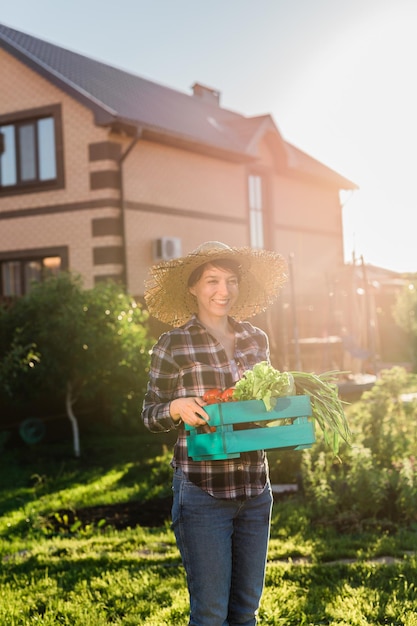 Fleißige junge Gärtnerin mit Strohhut holt an sonnigen Sommertagen ihre Erntekiste mit Tomaten ab Konzept des ökologischen Landbaus und des Gemüseanbaus