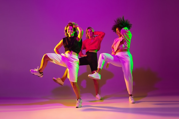 Flexibel. Sportliche Mädchen tanzen Hip-Hop in stilvoller Kleidung auf lila-rosa Hintergrund im Tanzsaal in grünem Neonlicht. Jugendkultur, Bewegung, Stil und Mode, Action. Modisches Porträt.