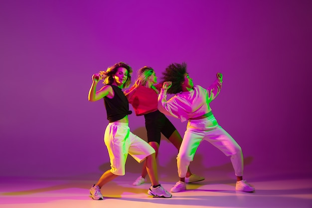 Flexibel. Sportliche Mädchen tanzen Hip-Hop in stilvoller Kleidung auf lila-rosa Hintergrund im Tanzsaal in grünem Neonlicht. Jugendkultur, Bewegung, Stil und Mode, Action. Modisches Porträt.