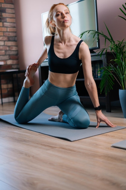 Flexible rothaarige junge Frau, die trainiert, Dehnübungen auf Yogamatten macht, während sie Fitnessvideos online auf dem Laptop anschaut. Konzept des Sporttrainings einer rothaarigen Dame während der Quarantäne.