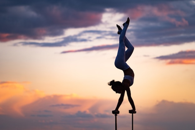 Flexible Zirkuskünstlerin hält das Gleichgewicht und macht Verrenkungen auf dem Dach gegen den dramatischen Sonnenuntergang und das Stadtbild. Motivation, Leidenschaft und Leistungskonzept.