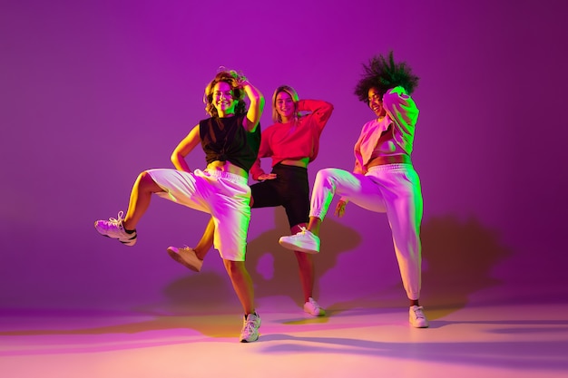 Foto flexível. meninas esportivas dançando hip-hop em roupas elegantes em fundo rosa-púrpura no salão de dança em luz de néon verde. cultura jovem, movimento, estilo e moda, ação. retrato elegante.