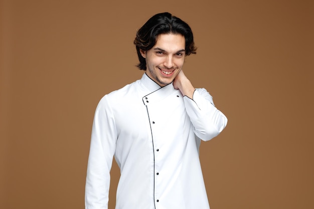 Flirty junger männlicher Koch in Uniform, der die Hand am Hals hält und isoliert auf braunem Hintergrund in die Kamera blickt