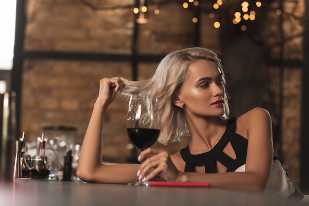 Flirty Stimmung. Attraktive blonde Frau, die an der Bartheke sitzt, Wein trinkt und mit jemandem über die Bar flirtet