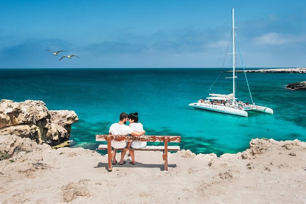 Flitterwochen-Reisendepaar umarmt auf einer Holzbank und genießt ihren tropischen Urlaub. Hochzeitsreise. Junges glückliches Paar, das sich am Strand zurücklehnt. Rückansicht eines verliebten Paares im Urlaub. Reisende