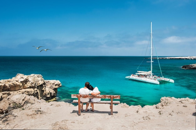 Flitterwochen-Reisendepaar umarmt auf einer Holzbank und genießt ihren tropischen Urlaub. Hochzeitsreise. Junges glückliches Paar, das sich am Strand zurücklehnt. Rückansicht eines verliebten Paares im Urlaub. Reisende