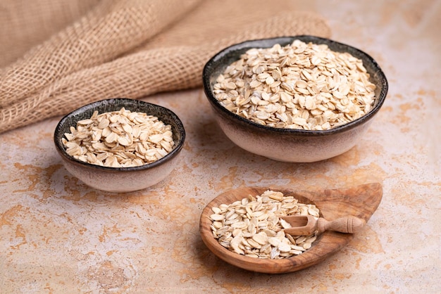 Flocos de aveia em tigelas de cerâmica feitas à mão e em uma tigela de madeira em serapilheira em uma colher de cereal de mesa de pedra bege