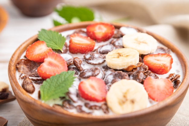 Flocos de milho de chocolate com leite e morango em uma tigela de madeira na superfície de madeira branca e têxteis de linho