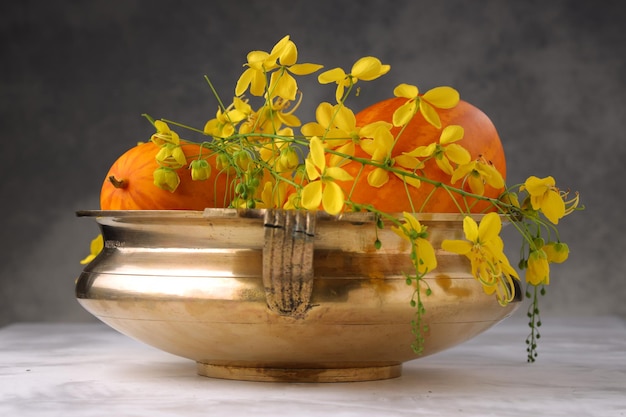 Flor de chuva dourada e pepino amarelo dispostos em um vaso tradicional de latão ou urule colocado no fundo texturizado branco