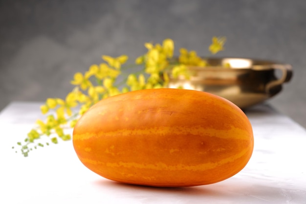 Flor de chuva dourada e pepino amarelo dispostos em uma superfície branca com fundo texturizado cinza e um vaso tradicional de latão ou urule disposto no fundo