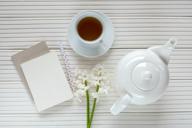 Flor de jacinto blanco, taza de té, tetera blanca y cuaderno en blanco sobre un fondo blanco de tablero de madera