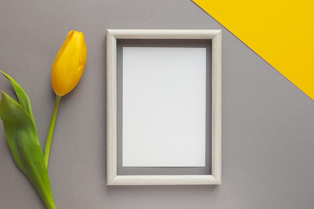 Flor de tulipán amarillo y papel vacío con marco de madera en mesa geométrica amarilla y gris