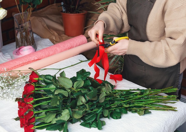 Floreria en el trabajo closeup manos femeninas cortan una cinta de color mujer haciendo un ramo de rosas rojas para