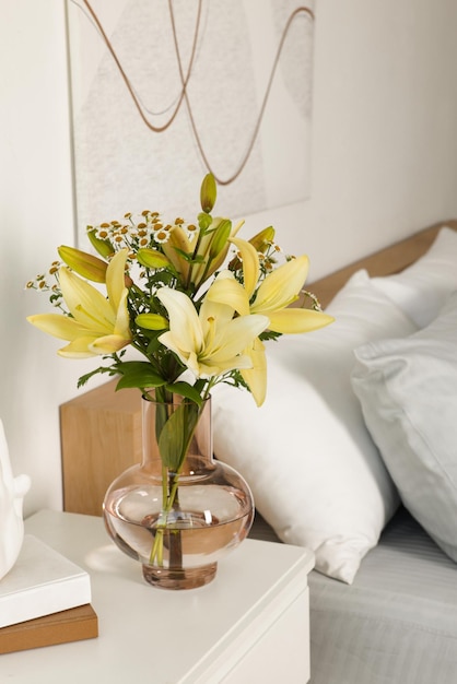 Florero con ramo de flores frescas en la mesita de noche blanca en el dormitorio