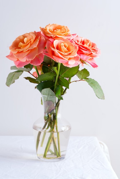 Florero de vidrio con un hermoso ramo de flores frescas rosas naturales en la mesa cubierta de tela blanca contra la pared gris claro.