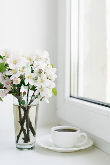 Flores da primavera em um vaso e uma xícara de chá ou café em uma caneca de porcelana branca no parapeito da janela