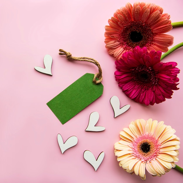 Foto flores gerbera, corações de madeira e etiqueta de papel verde em um fundo rosa pastel. vista do topo