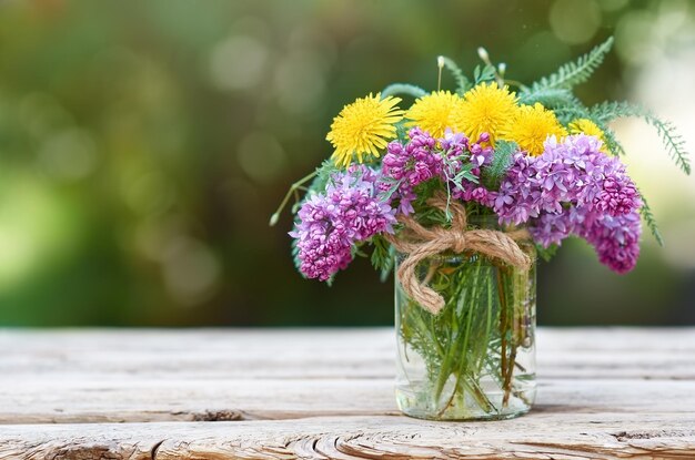 Flores lilas y dientes de león en frasco de vidrio sobre una mesa de madera frente a un fondo borroso