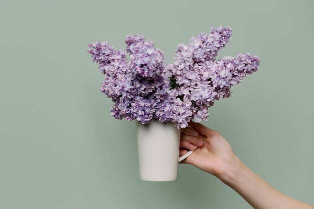 Flores lilás em copo branco nas mãos de mulher sobre fundo verde. Primavera, beleza, perfumaria, cosmetologia, férias, presente, conceito de decoração