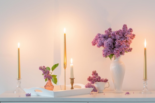 Flores lilás em vaso branco e velas douradas na parede branca de fundo