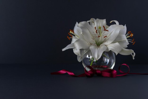 Flores de lirios blancos en un jarrón de vidrio con una cinta roja sobre un fondo oscuro. Decoración de interiores.