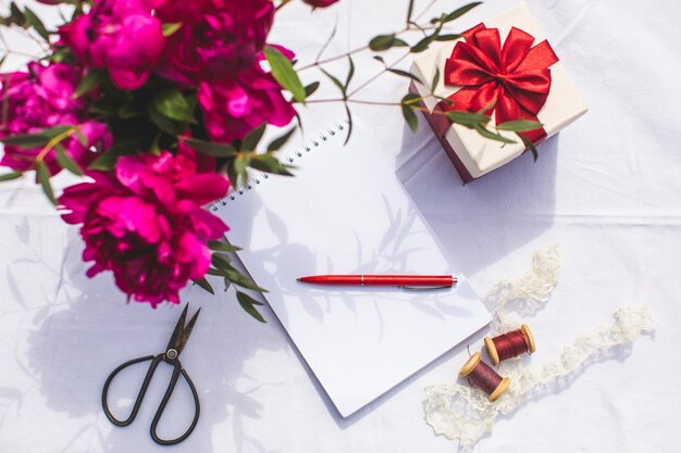 Flores de peonía y una pequeña caja de regalo colocada cerca de un bloc de notas abierto y un bolígrafo Escribiendo saludos en el cumpleaños o el día de la boda Sobrecarga sobre fondo blanco