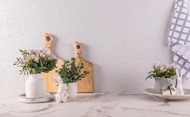 Flores de primavera en jarrón y jarra en la encimera de la cocina con decoraciones de Pascua, tablas de cortar, toallas, figuras de conejitos de Pascua