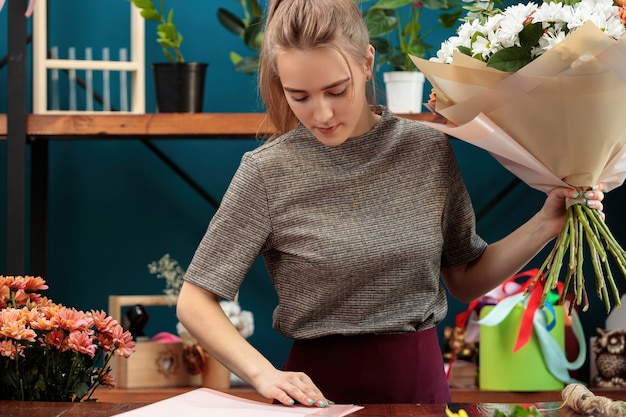 Florist macht einen Strauß. Ein junges erwachsenes Mädchen hält einen großen Strauß bunter Chrysanthemen in den Händen und wählt Papier zur Dekoration.