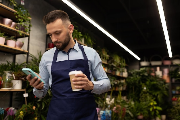 Florista de avental olha para o telefone com um copo de café nas mãos em uma floricultura