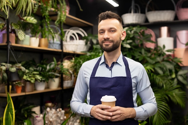 Un florista con un delantal azul y una taza de café en las manos se encuentra en una tienda de flores y ramos y